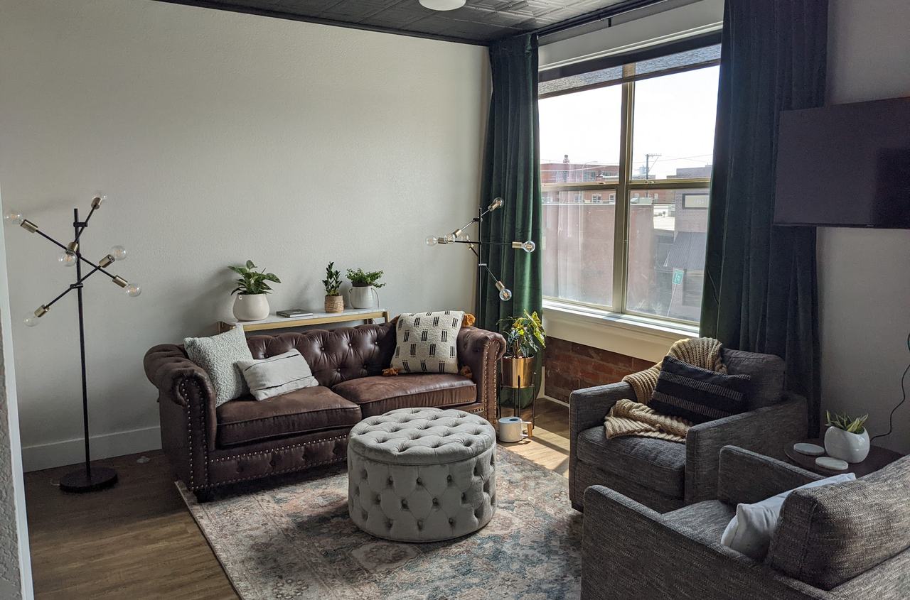 A finished lounge with a tufted leather sofa, a round tufted ottoman, two tweed armchairs, deep green velvet drapes at a tall window, and a dark pressed-tin ceiling.