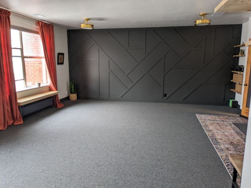 An open studio room with a charcoal geometric paneled accent wall, brass ceiling fixtures, rust-red curtains, and a window bench.
