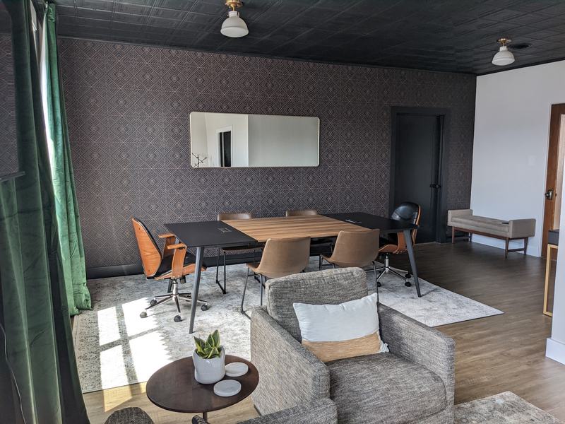 A conference room with geometric patterned wallpaper, a wood-and-black table with leather chairs, a long mirror, and a tweed armchair in the foreground.